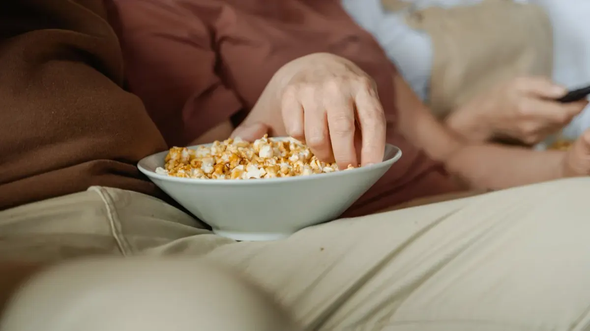 A dog sitting attentively beside an owner holding a bowl of popcorn on the couch
