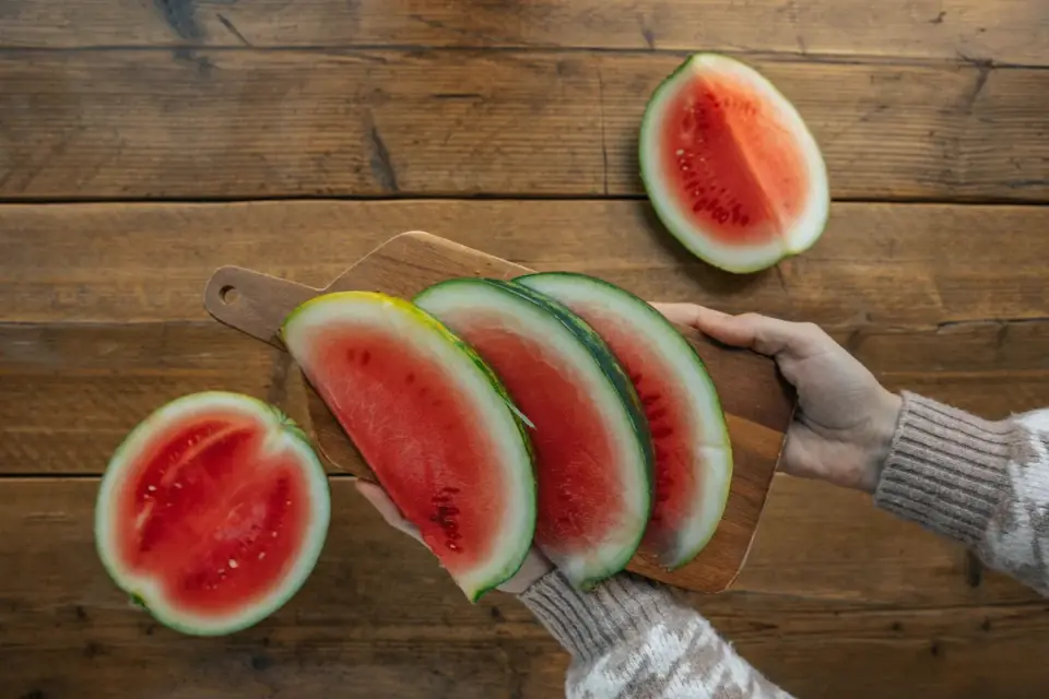 Hands trimming the tough green rind away from watermelon flesh on a cutting board
